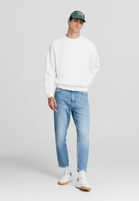 Felpa bianca oversize, jeans di jeans azzurro chiaro, berretto da baseball verde, sneakers bianche con dettagli verdi e calzini bianchi testurizzati.