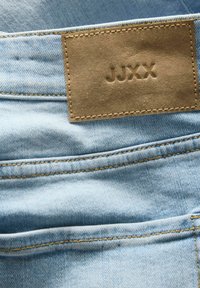 Vaqueros de mezclilla azul claro con costuras visibles y una etiqueta de cuero marrón con el grabado "JUXX". Presentan un diseño en el bolsillo trasero.