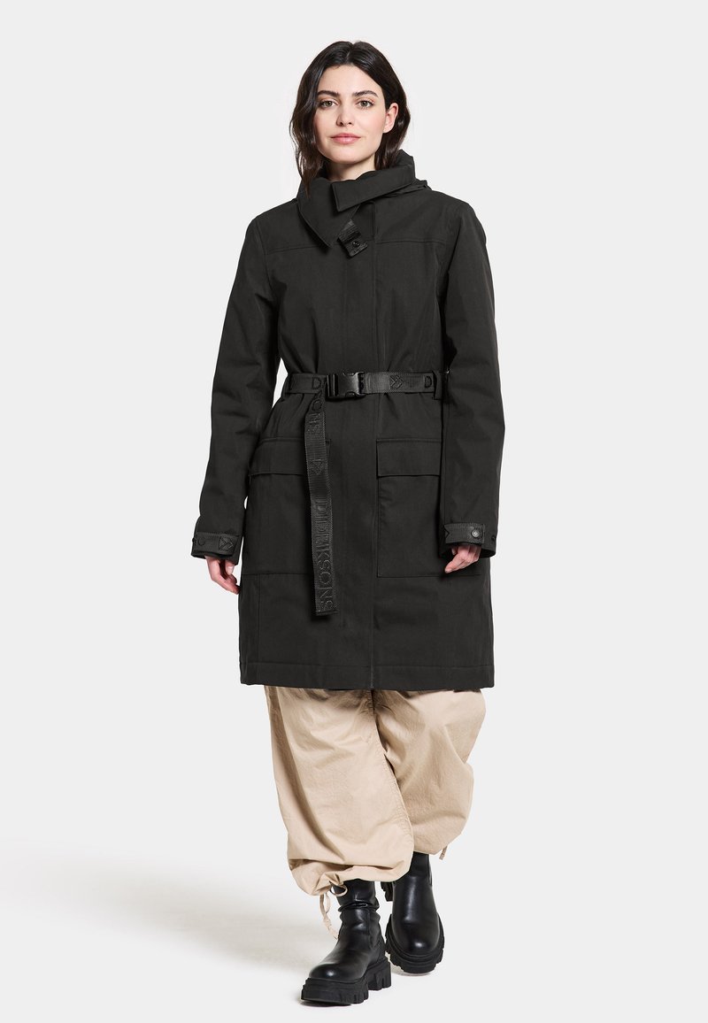 Schwarzer langer Mantel mit tailliertem Gürtel, hohem Kragen und Knopfakzenten. Verfügt über große Taschen und eine glatte, matte Textur. Kombiniert mit beigen Hosen.
