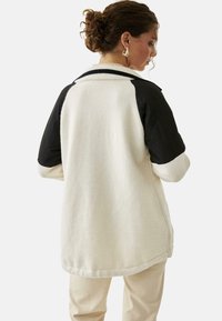 Chaqueta de felpa de gran tamaño en color crema con mangas acolchadas en negro. Cuello acanalado, hombros caídos y dobladillo curvado. Textura suave, diseño informal.