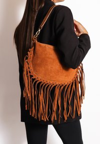Persoon in zwarte jas draagt een grote bruine suede schoudertas met lange franje op een witte achtergrond.