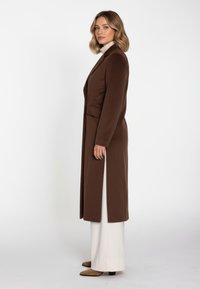 Lang brun frakke med en struktureret silhuet, lommer og sideslidser. Bærres over hvide bukser og brune støvler med hæl, og har et tekstureret stof.