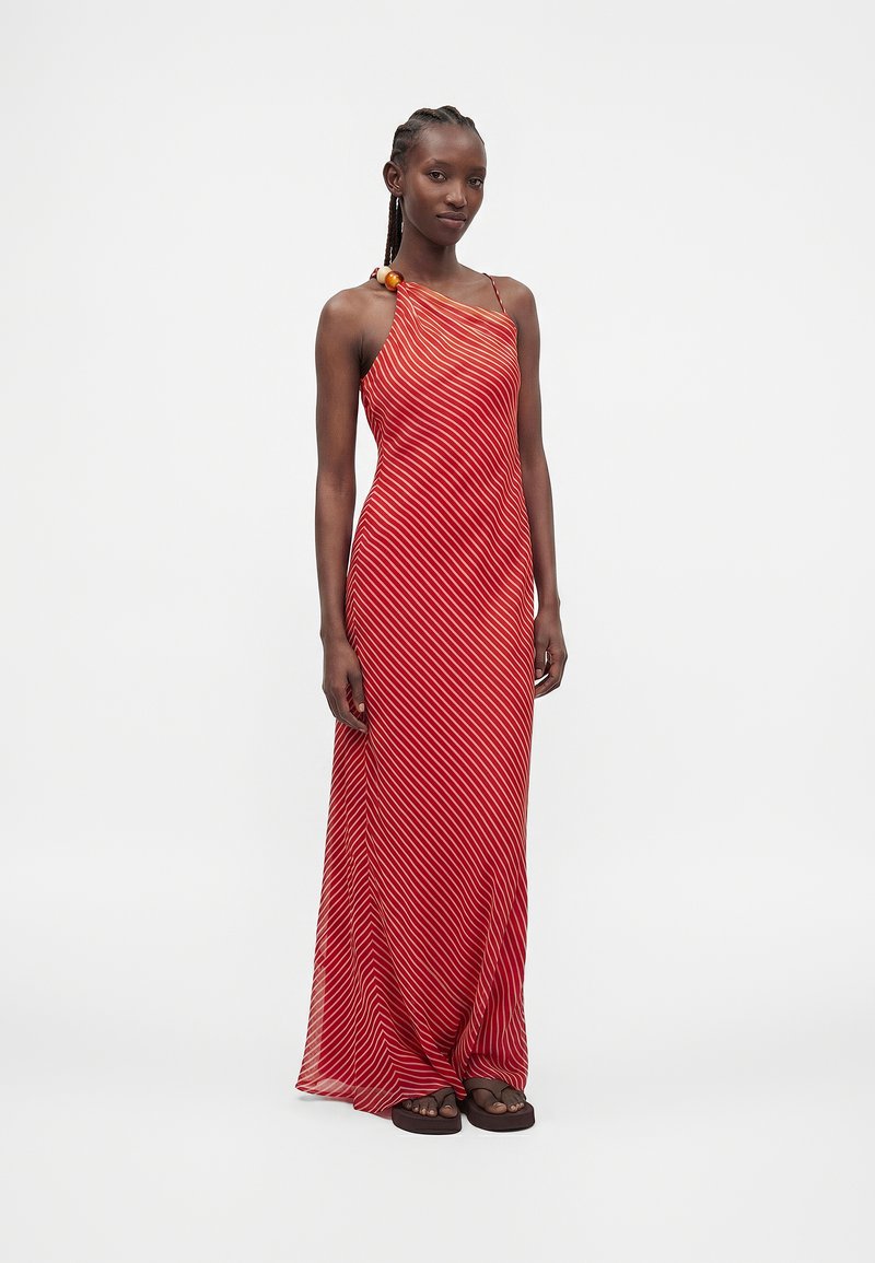 Femme portant une robe longue asymétrique à rayures diagonales rouges et blanches avec un détail en perles de bois et des sandales noires, debout devant un fond uni.