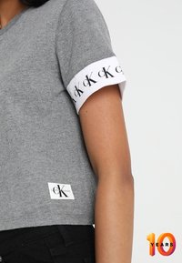 Šedá košile s krátkým rukávem a bílým manžetovým lemem, na které se opakuje logo "cK", nošena s černými kalhotami; malá obdélníková nášivka Calvin Klein blízko lemu.