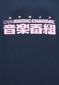 Υφασμα σε ναυτικό μπλε με ροζ κείμενο που γράφει "EDWIN music CHANNEL" και μεγάλοι ιαπωνικοί χαρακτήρες από κάτω, τοποθετημένοι στο κέντρο της επιφάνειας.