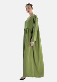 Lange, loszittende jurk in olijfgroen, met lange mouwen en een geplooide onderste sectie. Gemaakt van zachte stof met een gladde textuur.