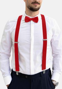 Punavärvilised kangast rihmad hõbedaste klambritega, koos valge nööpidega särk ja punane lips, kantud tumedate pükste seljas.