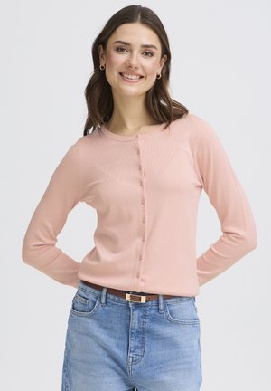 Noor naeratav naine, kes kannab heleroosat nööbitavat kardigani, siniseid teksaseid ja pruunikat vööd, seisab lihtsa heleda tausta ees.