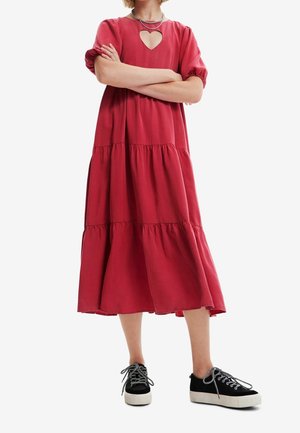 Persona vistiendo un vestido midi rojo suelto de capas con mangas abullonadas y un escote en forma de corazón en el pecho, combinado con zapatillas negras.