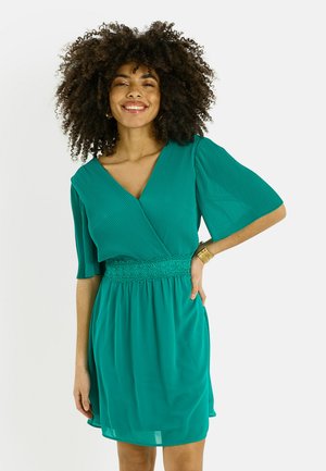 Mujer con cabello rizado sonriendo, vestida con un vestido de manga corta color azul verdoso con una cintura de encaje y una pulsera dorada, de pie frente a un fondo liso.