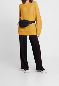 Žlutý pletený svetr s dlouhými rukávy, doplněný černými kalhotami s širokými nohavicemi a červenými pruhy po stranách a černou ledvinkou. Černé tenisky.