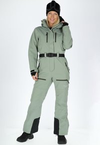 Green waterproof ski suit with a high collar, belt, zippered pockets, and black accents on cuffs and lower legs. Worn with a black beanie and goggles.
