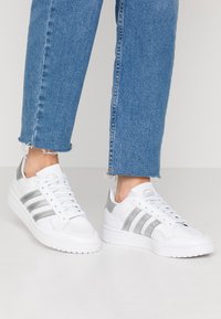 Vita sneakers med silverränder, slät syntetisk ovandel, rund tå och texturerad gummisula. Matchas med blå jeans med fransar.