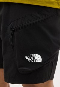 Μαύρο σορτς με τσεπάκι με φερμουάρ στο πλάι που φέρει το λογότυπο The North Face σε λευκό, φορεμένο με κίτρινη μπλούζα.