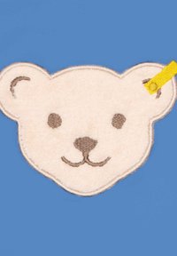Parche en forma de cara de oso suave y color crema con bordado marrón en los ojos y la nariz, que presenta una etiqueta de tela amarilla en la parte superior.