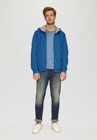 Blauwe hoodie met ritssluiting en grijze binnenvoering, gecombineerd met een lichtblauw T-shirt, donkere spijkerbroek en witte sneakers. Bevat twee voortassen.