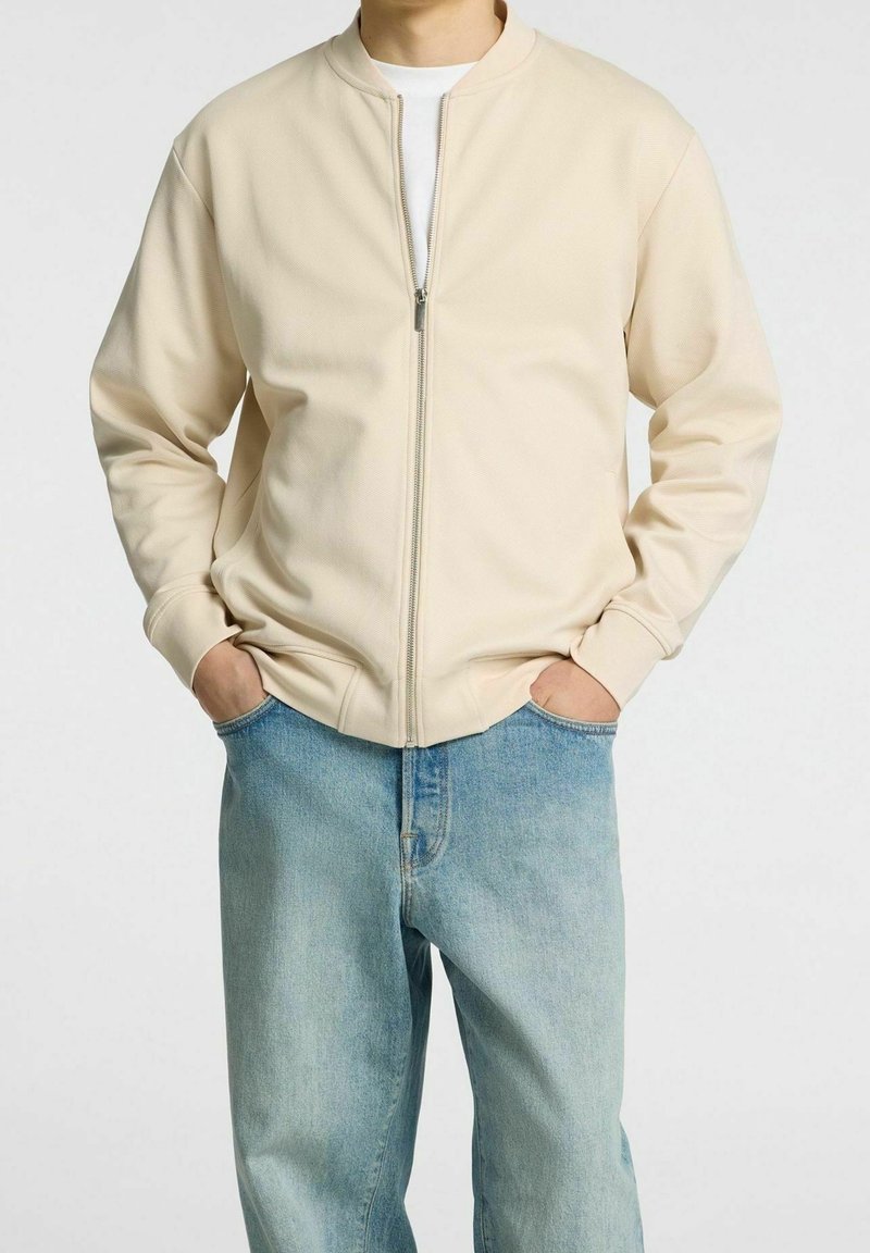 Hombre vistiendo una chaqueta ligera beige con cremallera sobre una camisa blanca, con las manos en los bolsillos de unos jeans de mezclilla azul claro, contra un fondo blanco.