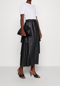 Donna che indossa un top bianco con maniche a sbuffo, una gonna midi in pelle nera a strati, mule nere con tacco e tiene in mano una borsa a tracolla in pelle nera.