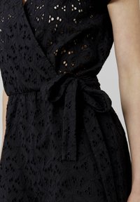 Robe portefeuille noire brodée avec des motifs floraux découpés, une ceinture à nouer à la taille et un tissu texturé. Vue en gros plan du design.
