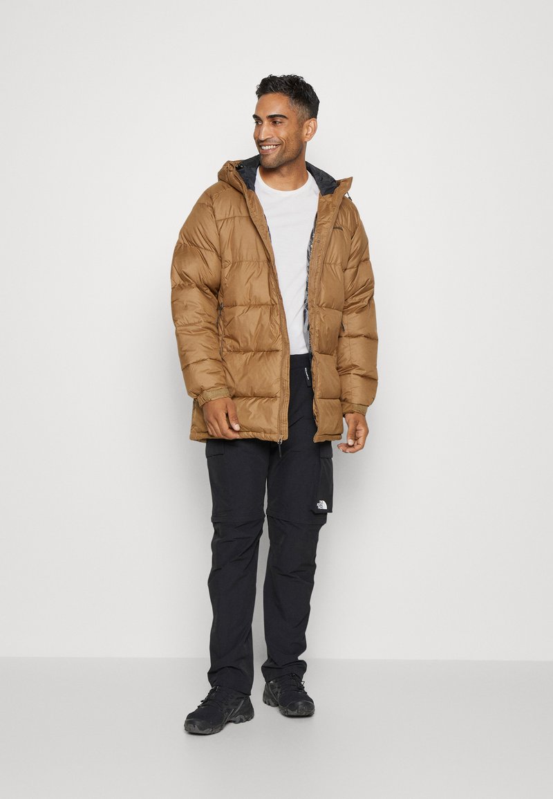 Mężczyzna w brązowej kurtce puchowej, noszący białą koszulę, czarne spodnie cargo i czarne trampki, stoi i uśmiecha się na jednolitym tle.