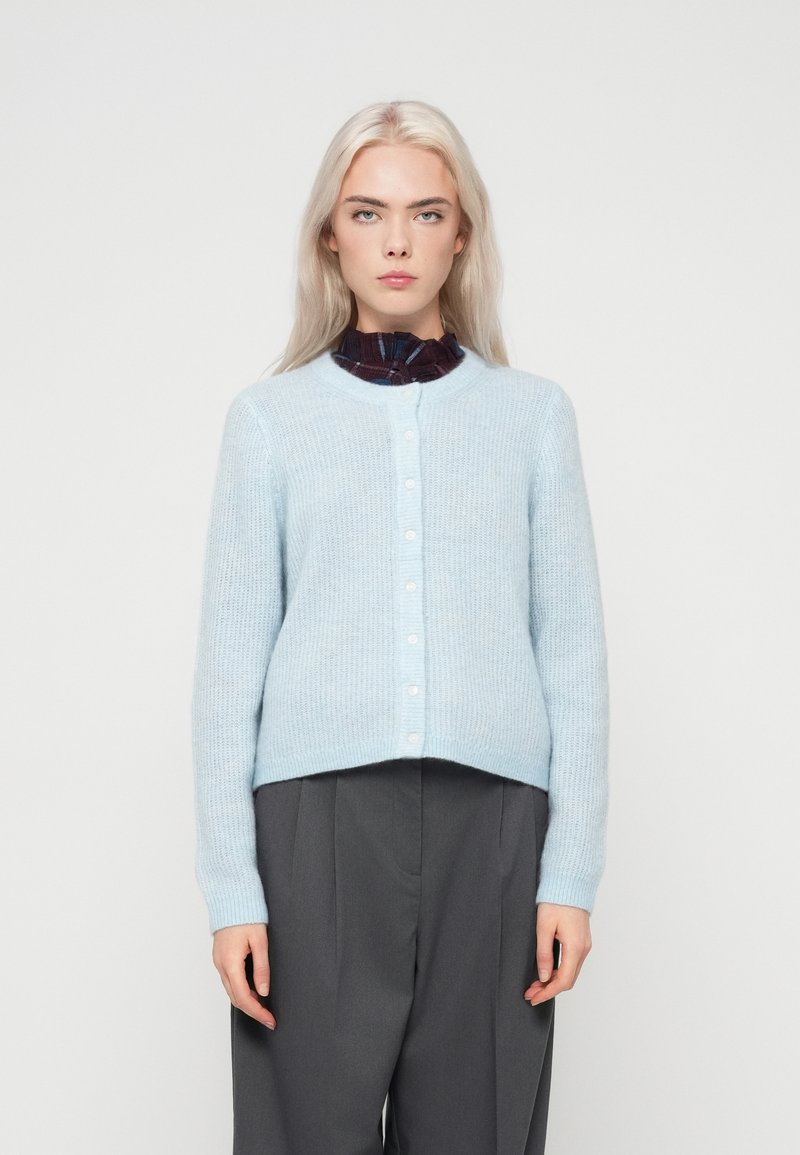 Blondynka w jasnoniebieskim zapinanym kardiganie noszącym się na ciemnej bluzce z falbanami, w szarych, wysokich szerokich spodniach.