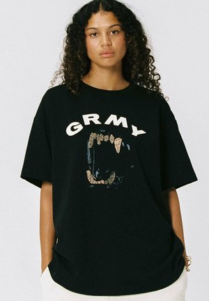 Mujer joven con cabello rizado que lleva una camiseta negra con el texto "GRMY" y un diseño de boca abierta con dientes visibles.
