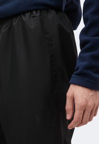 Άτομο που φοράει μπλε φλις μπλούζα και φαρδύ μαύρο παντελόνι, απεικονίζεται από τη μέση μέχρι τους μηρούς, με το δεξί χέρι χαλαρό δίπλα στο σώμα.
