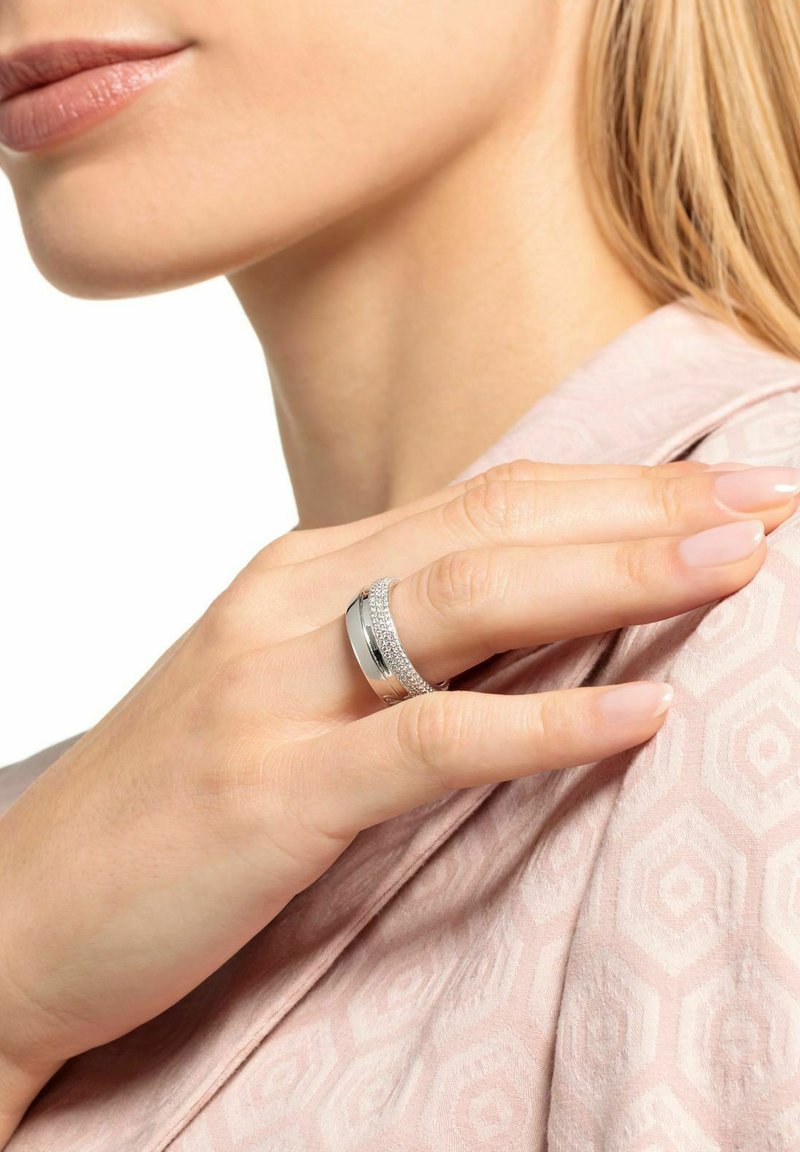 Nahaufnahme einer Hand mit natürlichen Nägeln, die einen silbernen Ring mit kleinen Diamanten trägt und auf einer Schulter liegt, die mit pinkfarbenem, gemustertem Stoff bekleidet ist.