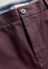 Pantaloni violet închis, cu o textură netedă, având un sistem de închidere cu un singur buton și un brâu cu un model contrastant.