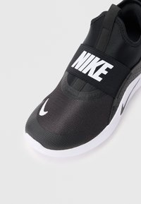 Baskets noires de sport avec une tige en mesh, sangle élastique arborant le logo "NIKE" et semelle blanche. Forme courbée et détails texturés sur le bout.