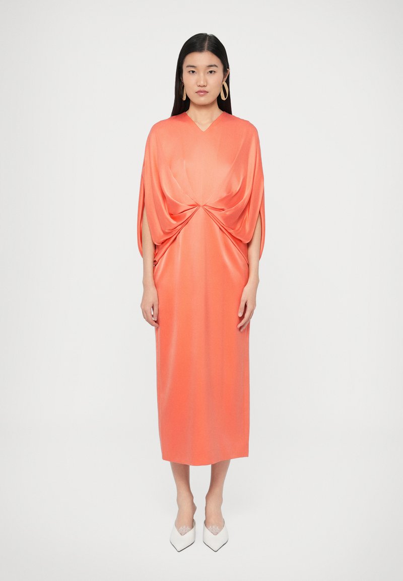 Φόρεμα κοραλλί σατέν με V-λαιμόκοψη και ρεβέρ μαν�ίκια, με στριφογυρισμένη λεπτομέρεια στο μπροστινό μέρος. Συνδυάζεται με άσπρα τακούνια μυτερής μύτης.