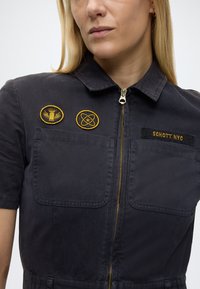 Femme portant une chemise zippée bleu marine foncé à manches courtes, avec deux poches poitrine, des écussons brodés jaunes et le texte "SCHOTT NYC".