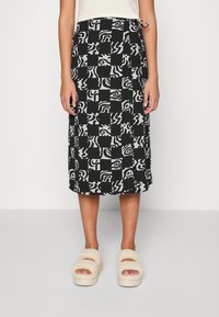 Saia midi em preto e branco com padrões abstratos, apresentando uma cintura com cordão e um corte reto. Usada com sandálias bejes fofas.