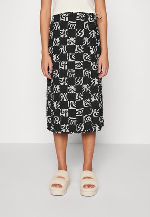 Saia midi em preto e branco com padrões abstratos, apresentando uma cintura com cordão e um corte reto. Usada com sandálias bejes fofas.