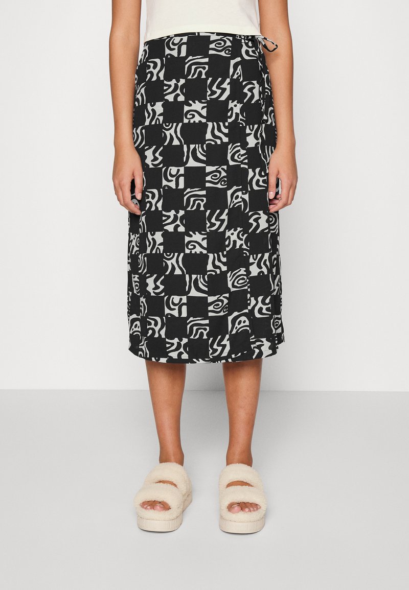 Saia midi em preto e branco com padrões abstratos, apresentando uma cintura com cordão e um corte reto. Usada com sandálias bejes fofas.