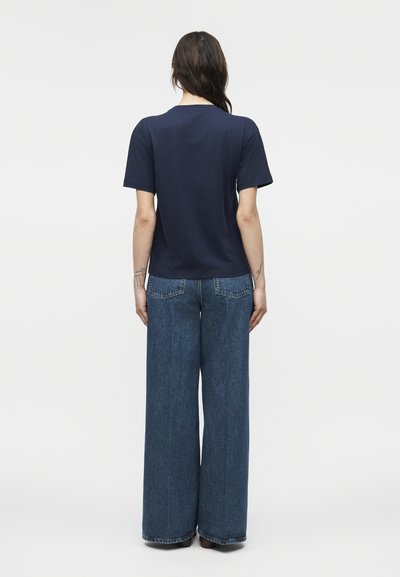 Γυναίκα στέκεται με την πλάτη στραμμένη φοράει μπλε σκούρο κοντομάνικο πουκάμισο και φαρδιά μπλε τζιν παντελόνια μπροστά σε απλό φόντο.