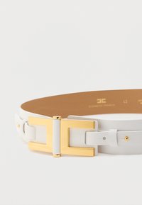 Λευκή δερμάτινη ζώνη με μεγάλη χρυσή ορθογώνια αγκράφα και διακοσμητικά χρυσά καρφιά, τοποθετημένη επίπεδα σε λευκή επιφάνεια.