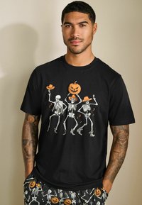 Sort bomulds t-shirt med et farverigt grafik af tre dansende skeletter med orange hatte, der holder en græskar. Designet inkluderer musiknoter.