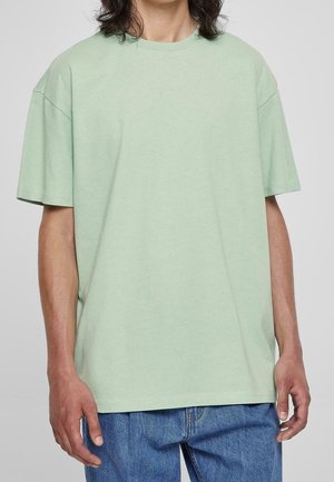 Person trägt ein lockeres hellgrünes kurzärmeliges T-Shirt und blaue Jeans, steht vor einem einfarbigen grauen Hintergrund.