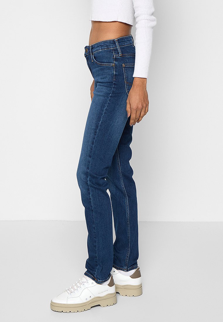 Jean en denim bleu skinny avec une taille mi-haute, poches avant et détails de couture minimalistes. Associé à des baskets blanches à plateforme.