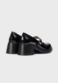 Zapatos de charol negros con tacón cuadrado y una correa que presenta una hebilla plateada. Diseño de punta redonda y textura suave.