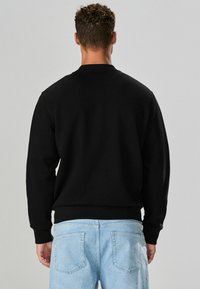 Felpa nera con collo a giro, maniche lunghe e polsini e orlo a coste. Indossata con jeans azzurri, mostra uno stile casual.