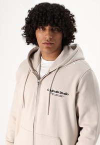 Beige ritshoodie gemaakt van zachte stof met een voorzak, met een logo op de borst. De trekkoorden hebben zilveren hardware-accenten.
