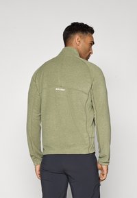 Giacca in pile verde con colletto alto, dotata di un logo sulla parte posteriore, cuciture piatte e maniche lunghe. Materiale testurizzato e design aderente.