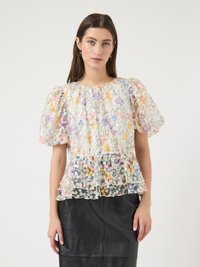 Vrouw die een doorschijnend bloesje met bloemenprint en pofmouwen met ruches aan de zoom draagt, ingestopt in een zwarte leren rok met hoge taille, staand tegen een effen achtergrond.