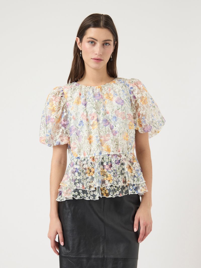 Vrouw die een doorschijnend bloesje met bloemenprint en pofmouwen met ruches aan de zoom draagt, ingestopt in een zwarte leren rok met hoge taille, staand tegen een effen achtergrond.