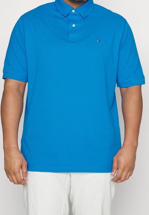 Mand iført en klar blå kortærmet poloshirt med krave og to knapper, kombineret med hvide bukser, stående mod en ensfarvet baggrund.