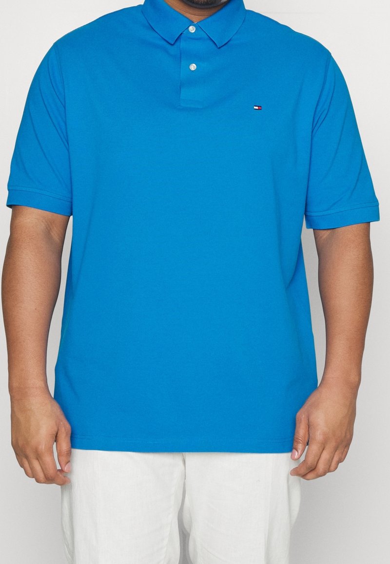 Homme portant un polo bleu vif à manches courtes avec col et deux boutons, associé à un pantalon blanc, debout devant un fond uni.