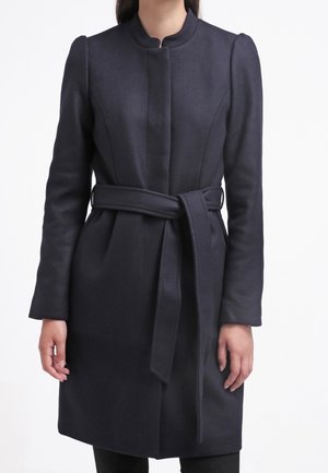 Frau trägt einen langen schwarzen Mantel mit Gürtel und hohem Kragen, steht vor einem weißen Hintergrund.