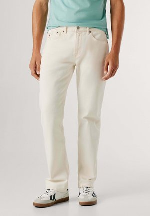 Hombre con vaqueros de pierna recta color blanco roto, zapatillas blancas con rayas negras y una camisa de color teal claro, de pie frente a un fondo liso.